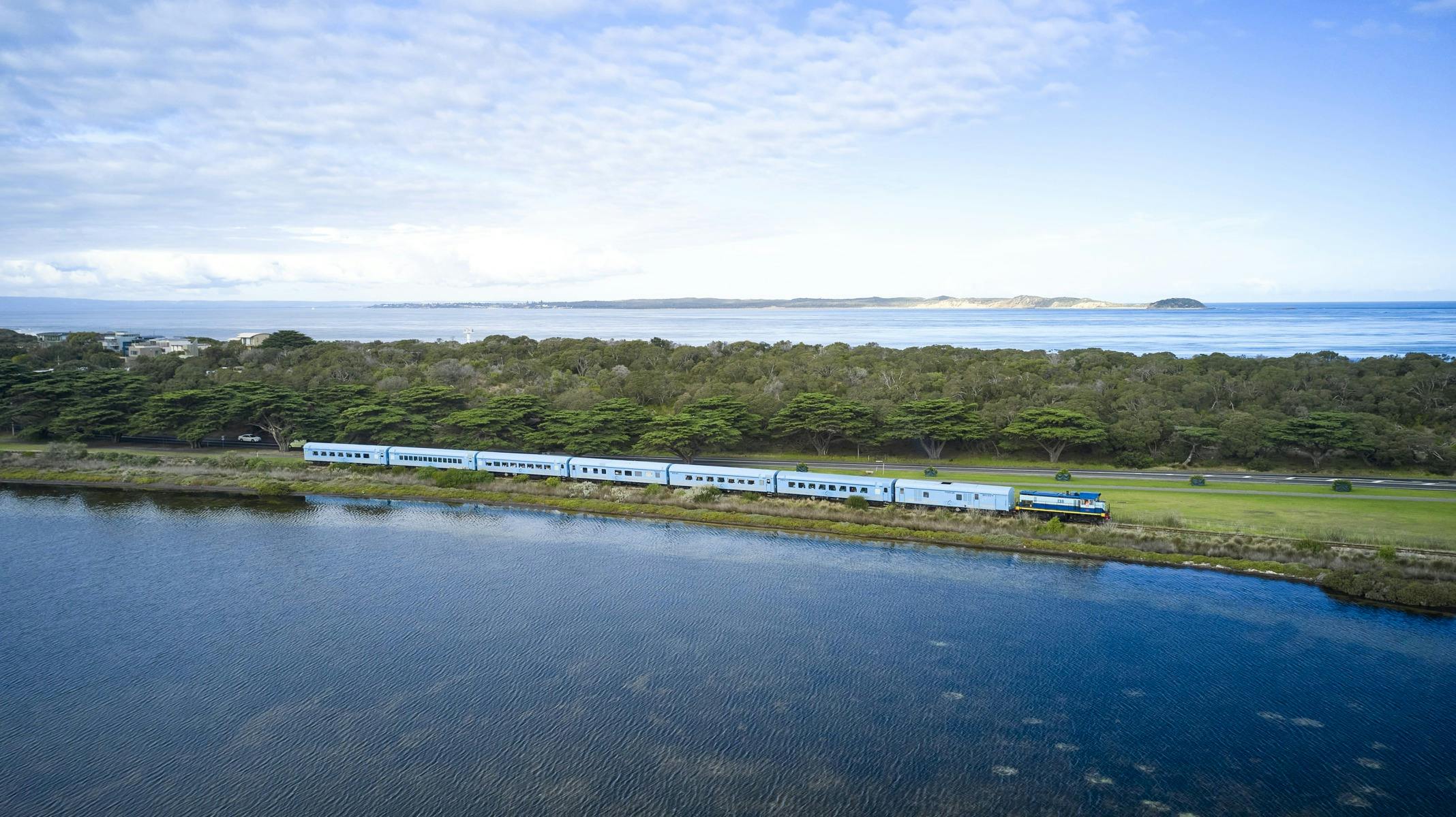 Blue train with 8 carriages and diesel locomotive passes by bay