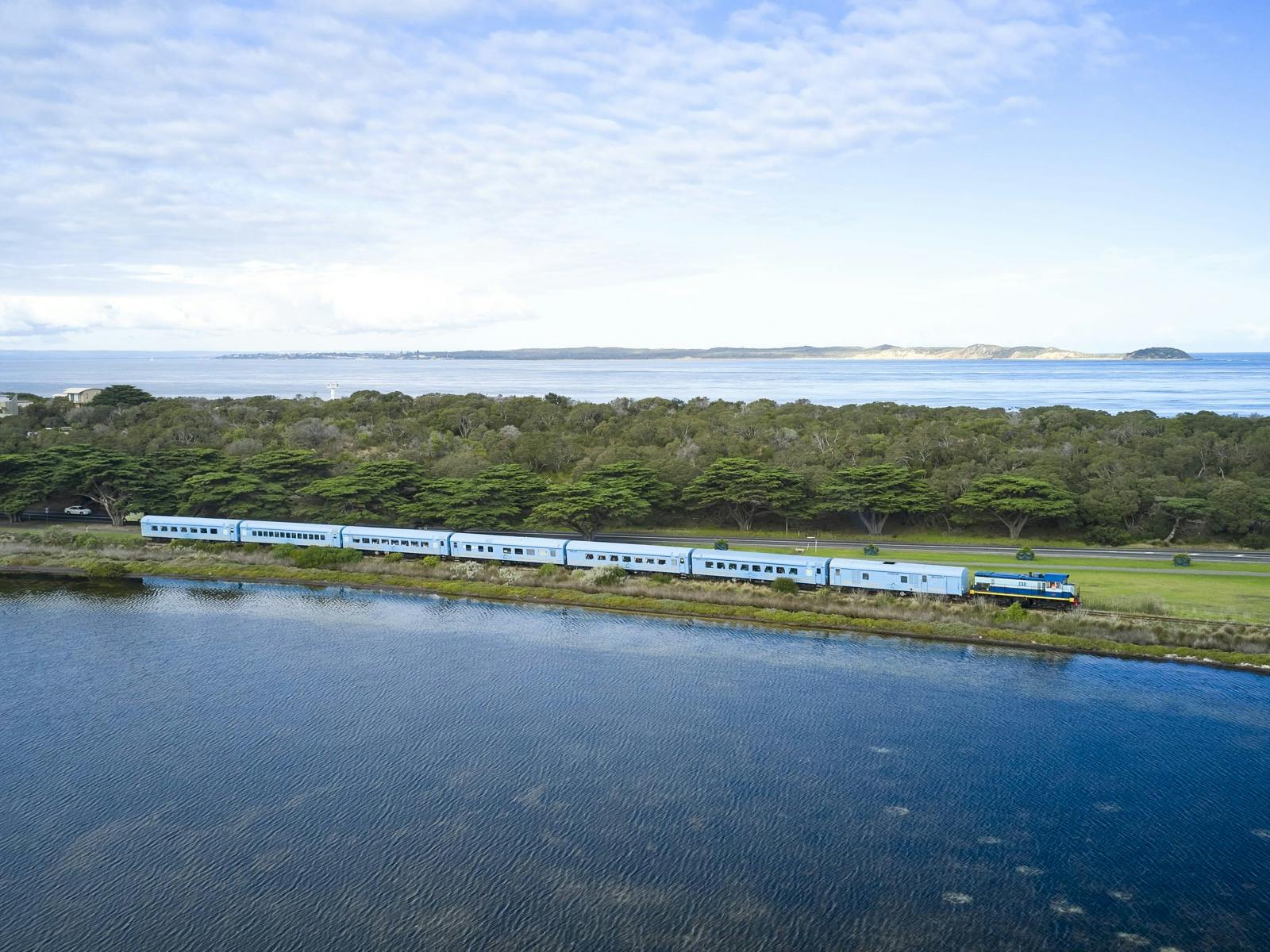 Blue train with 8 carriages and diesel locomotive passes by bay