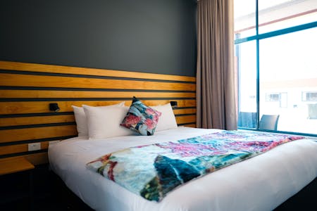 Double bed with white sheets and floral throw next to a window. Wooden head board on navy blue wall