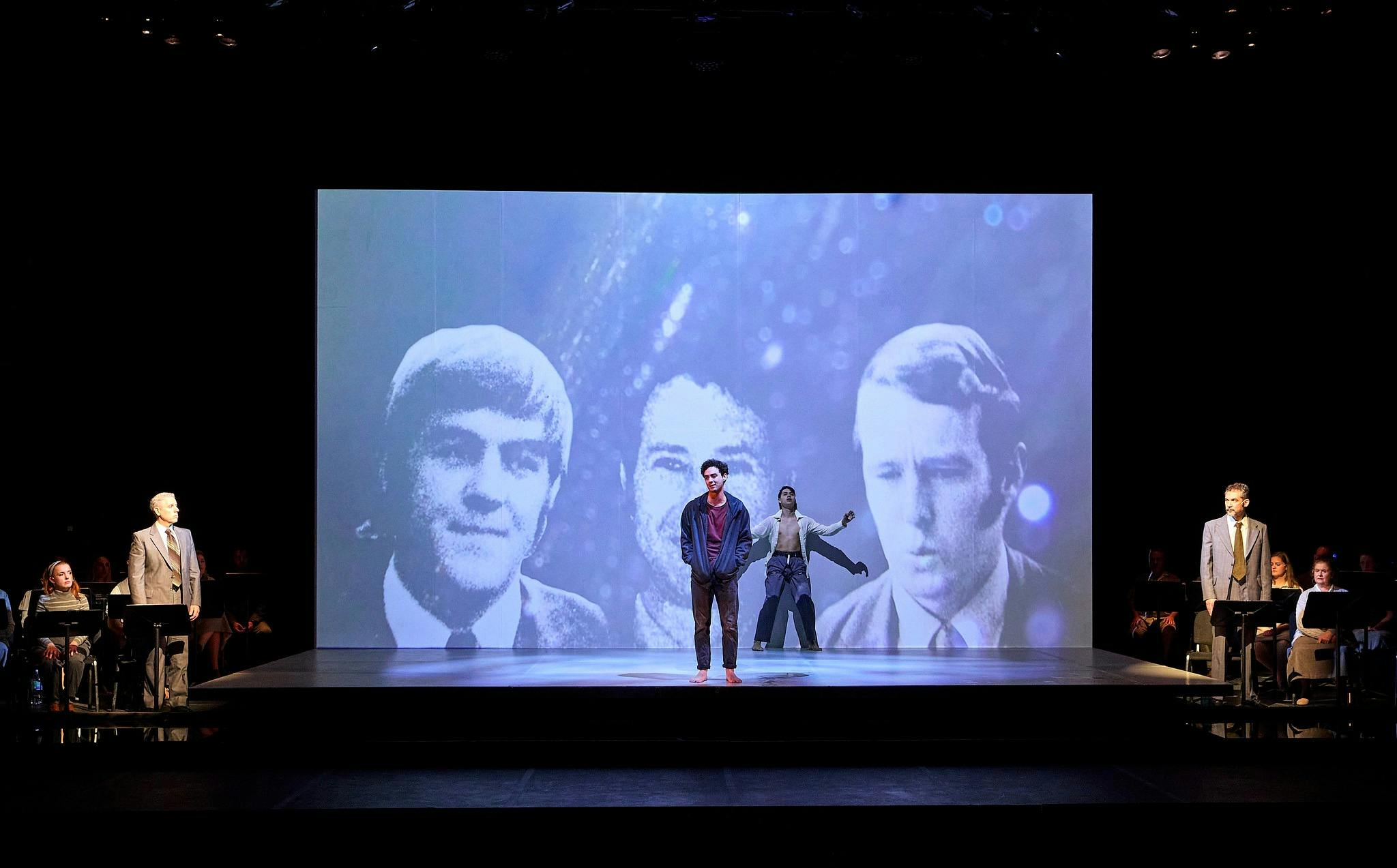 Performer stands before projected portraits as cast and musicians frame the stage.