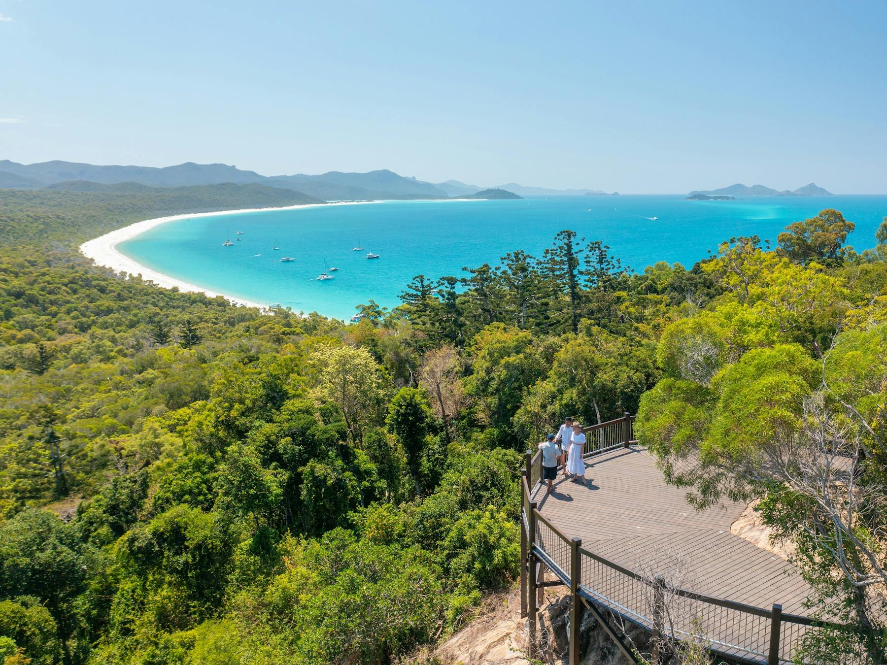 People on a hilltop lookout point overlooking the trees, long stretch of white sandy beach and ocean