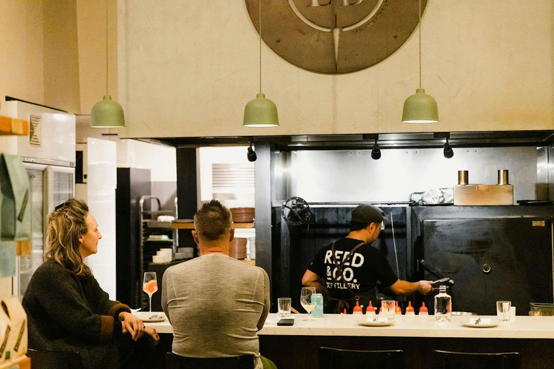 Two customers sit at a bar, watching a chef prepare their meals
