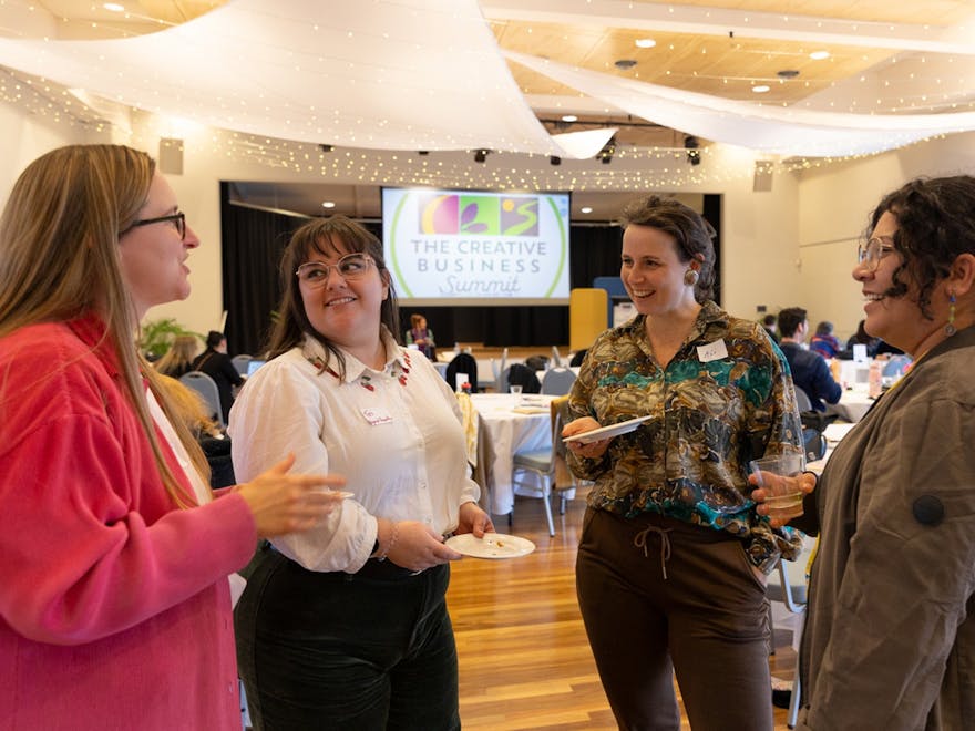 Four people standing eating lunch with the event stage in the background. Smiling & chatting