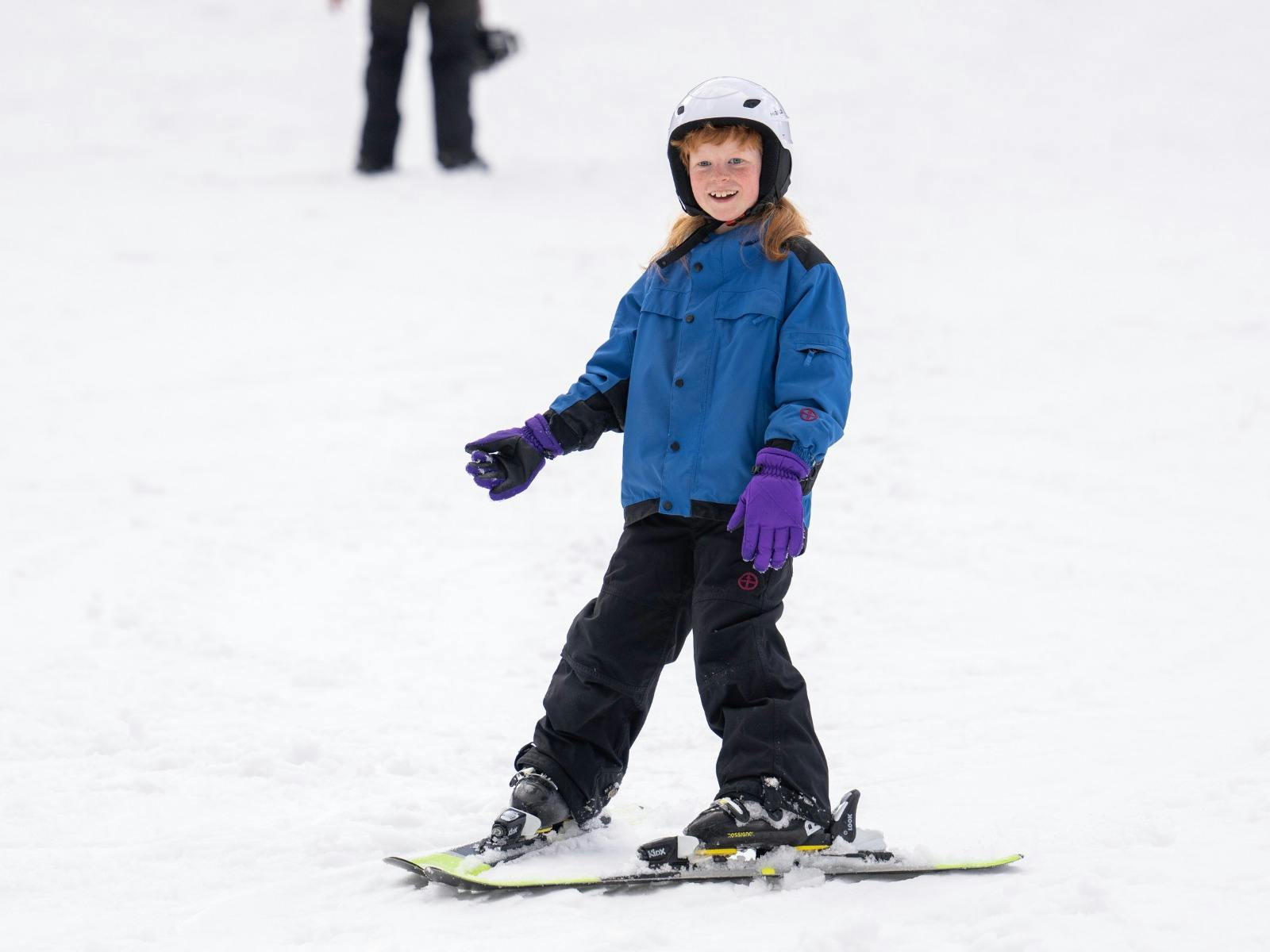 A boy skis on a gentle slope in a snowplough stance. The background is covered in snow.