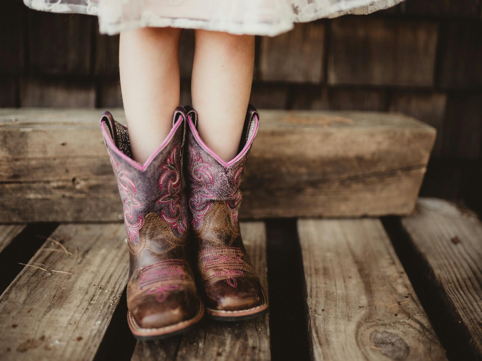 A close-up of brown and pink cowboy boots on the feet of a young girl, resting on a weathered wooden