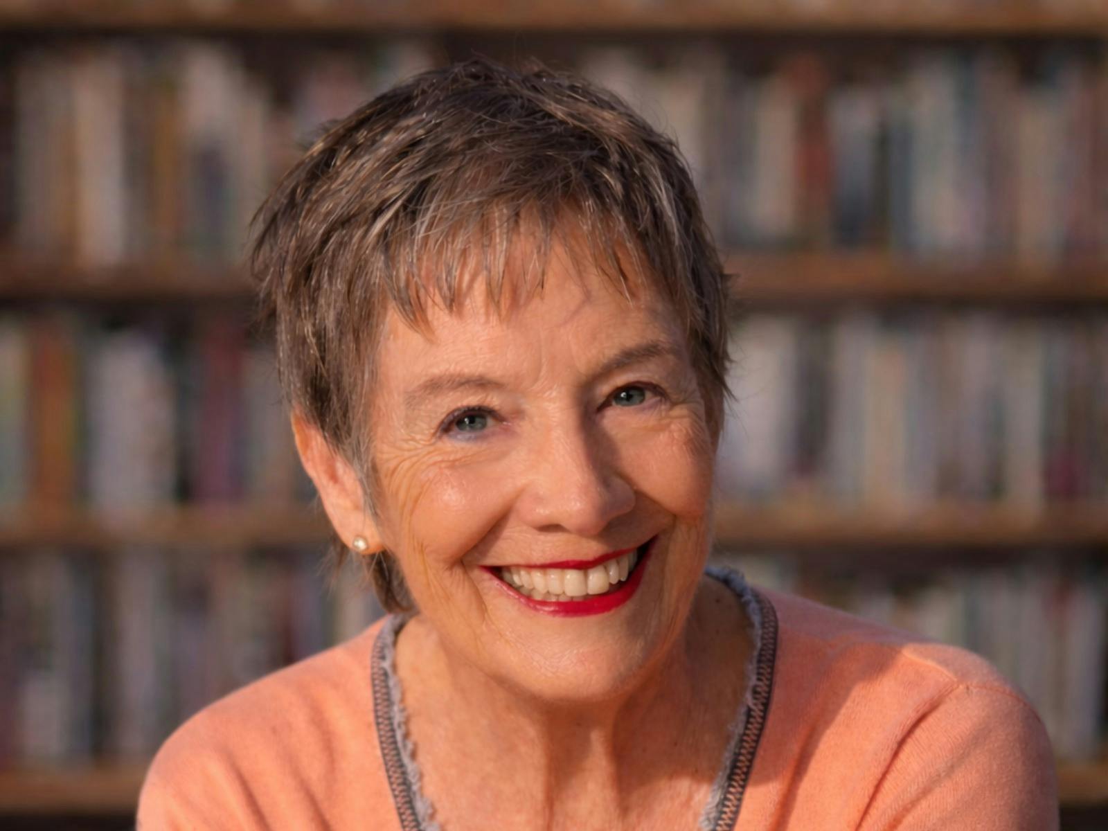A smiling, mature woman with short, gray hair, looking directly at the camera in front of a blurred