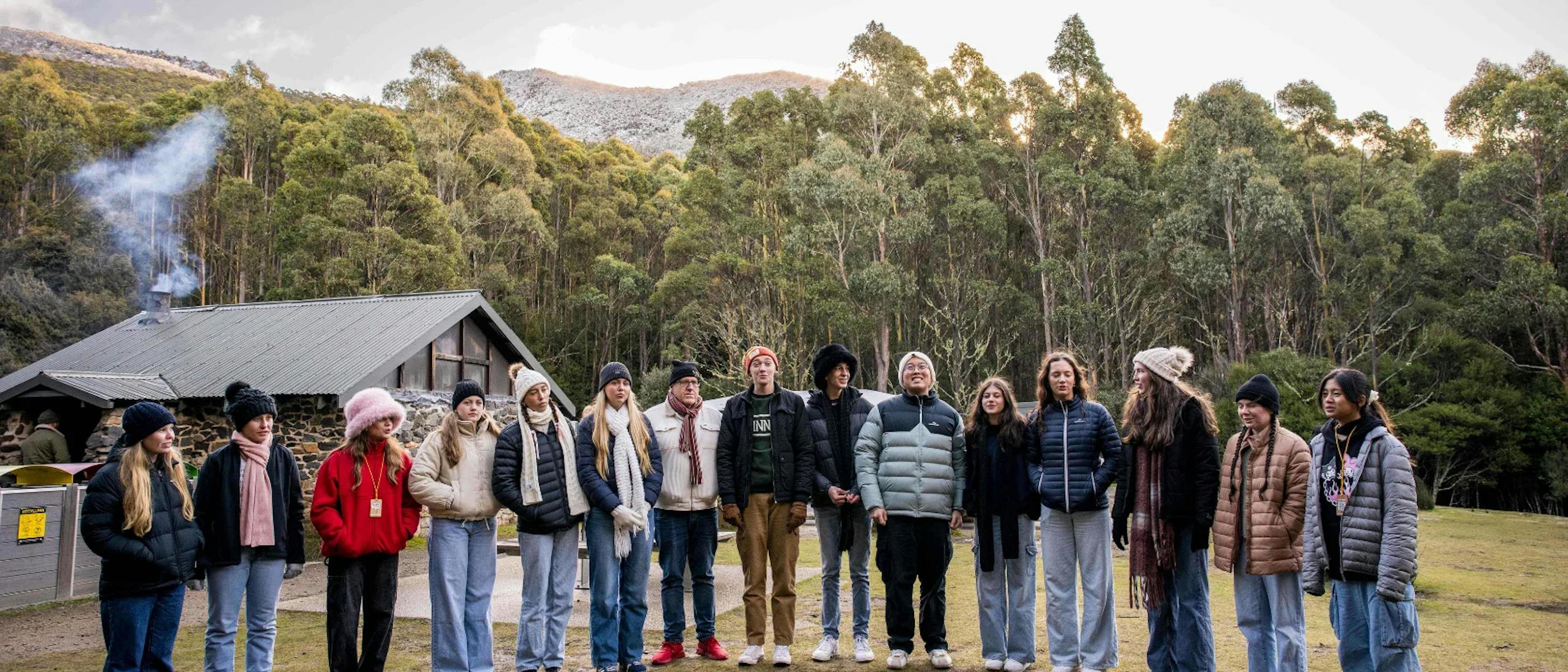 A group of people dressed in winter clothing stand outside a stone hut in a forested area