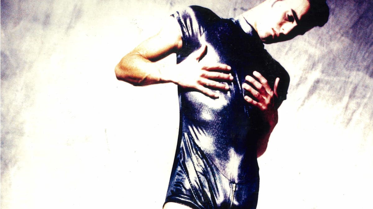 A person in a black leather look jumpsuit with short shorts poses with their hands on their chest