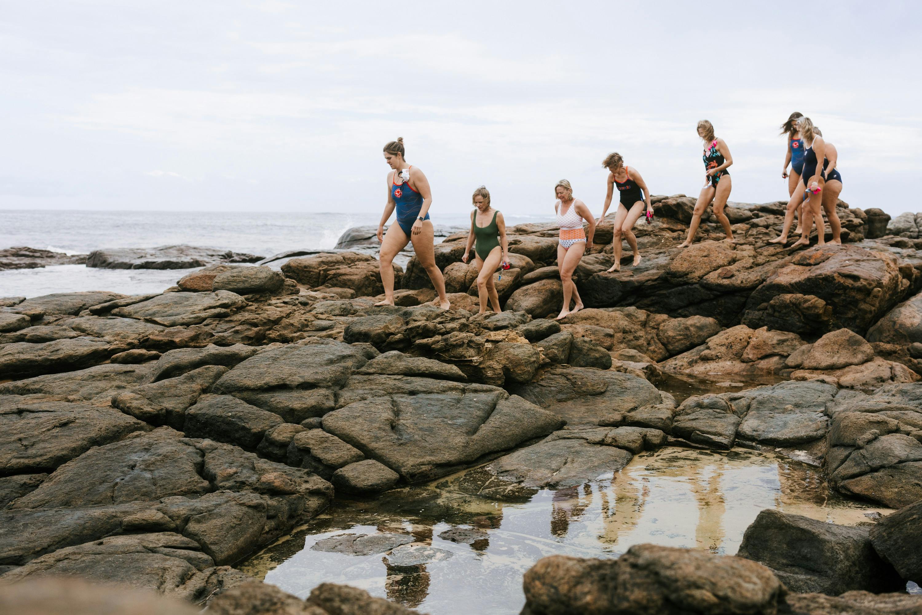 people in swim suits scramble over large granite rocks with rock pools in the foreground