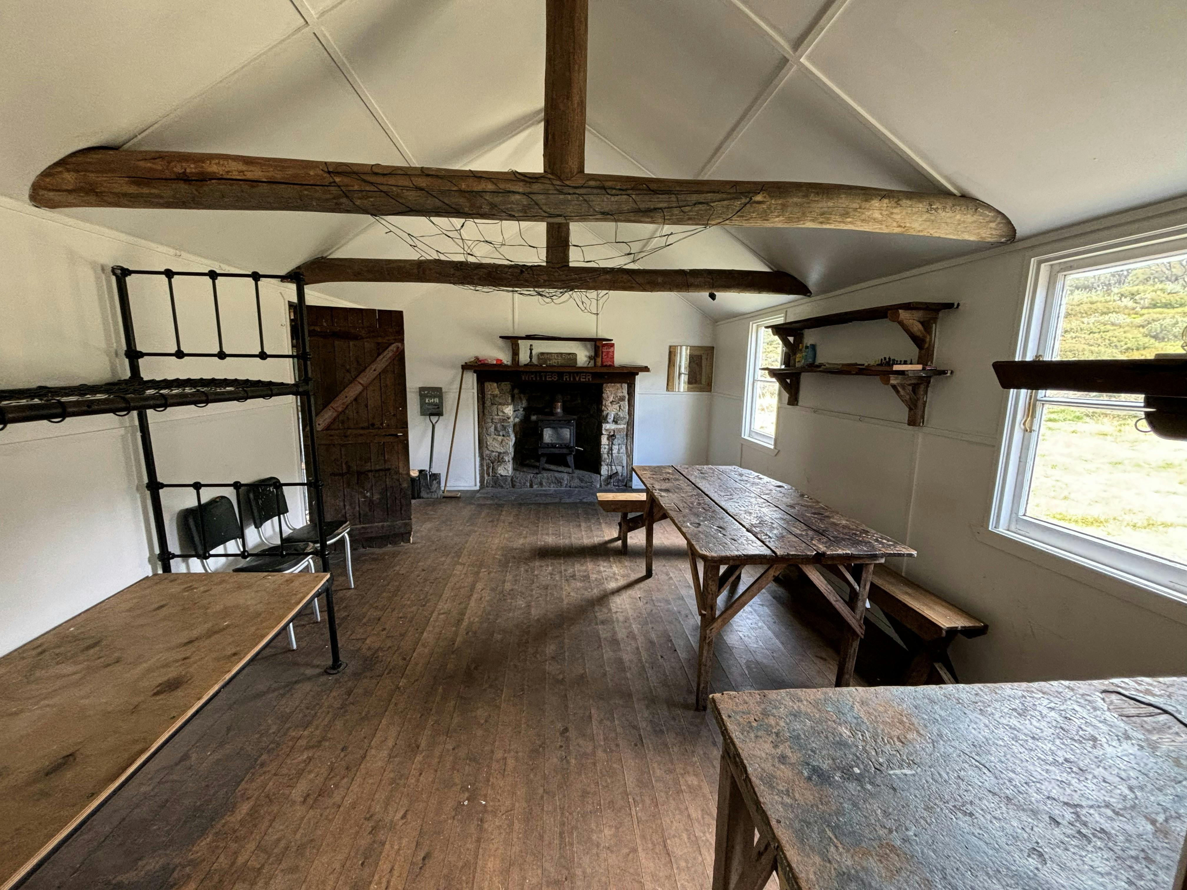 The interior of a timber and tin hut showing metal bunks, wooden tables and benches and a fireplace.