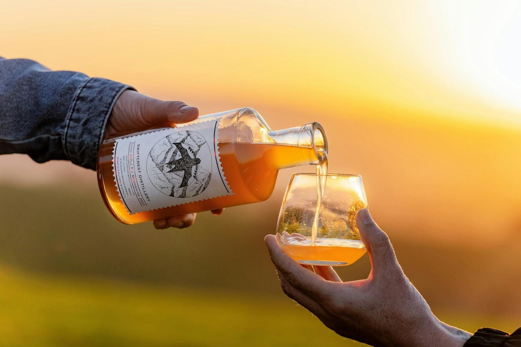 A hand pours amber-colored gin from a Swiftcrest Distillery bottle into a glass at sunset.