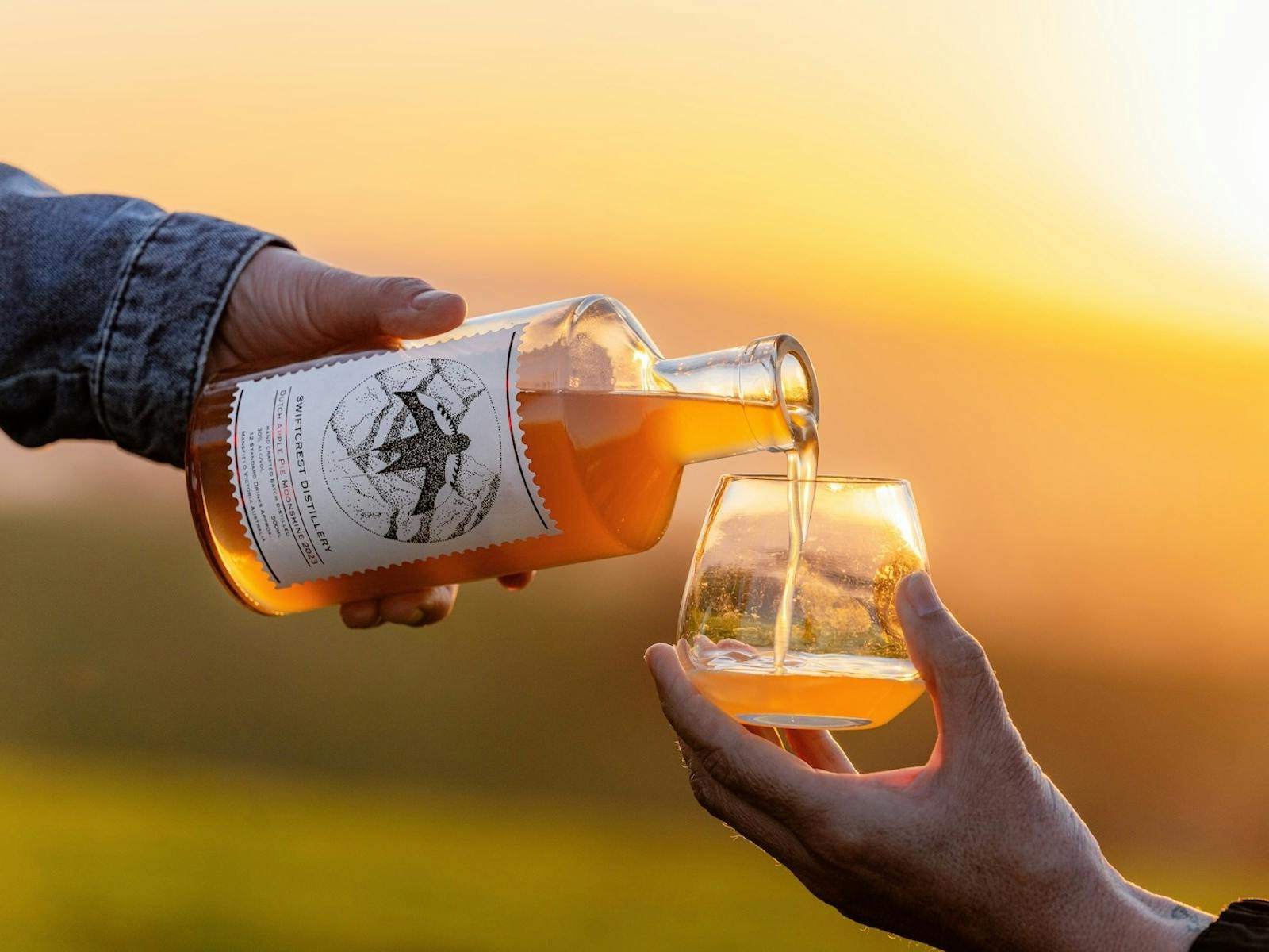 A hand pours amber-colored gin from a Swiftcrest Distillery bottle into a glass at sunset.