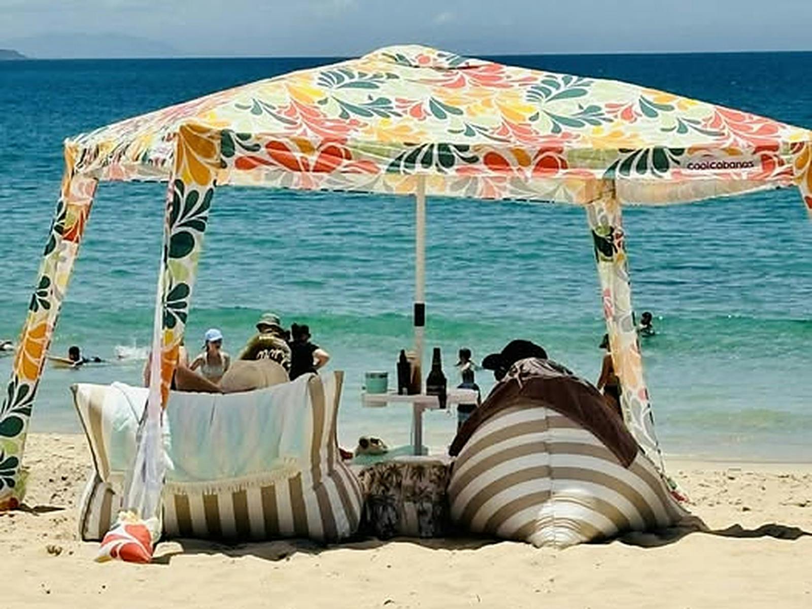 Beachgoers relaxing on striped beanbags under a floral beach cabana with a small drinks table, looki