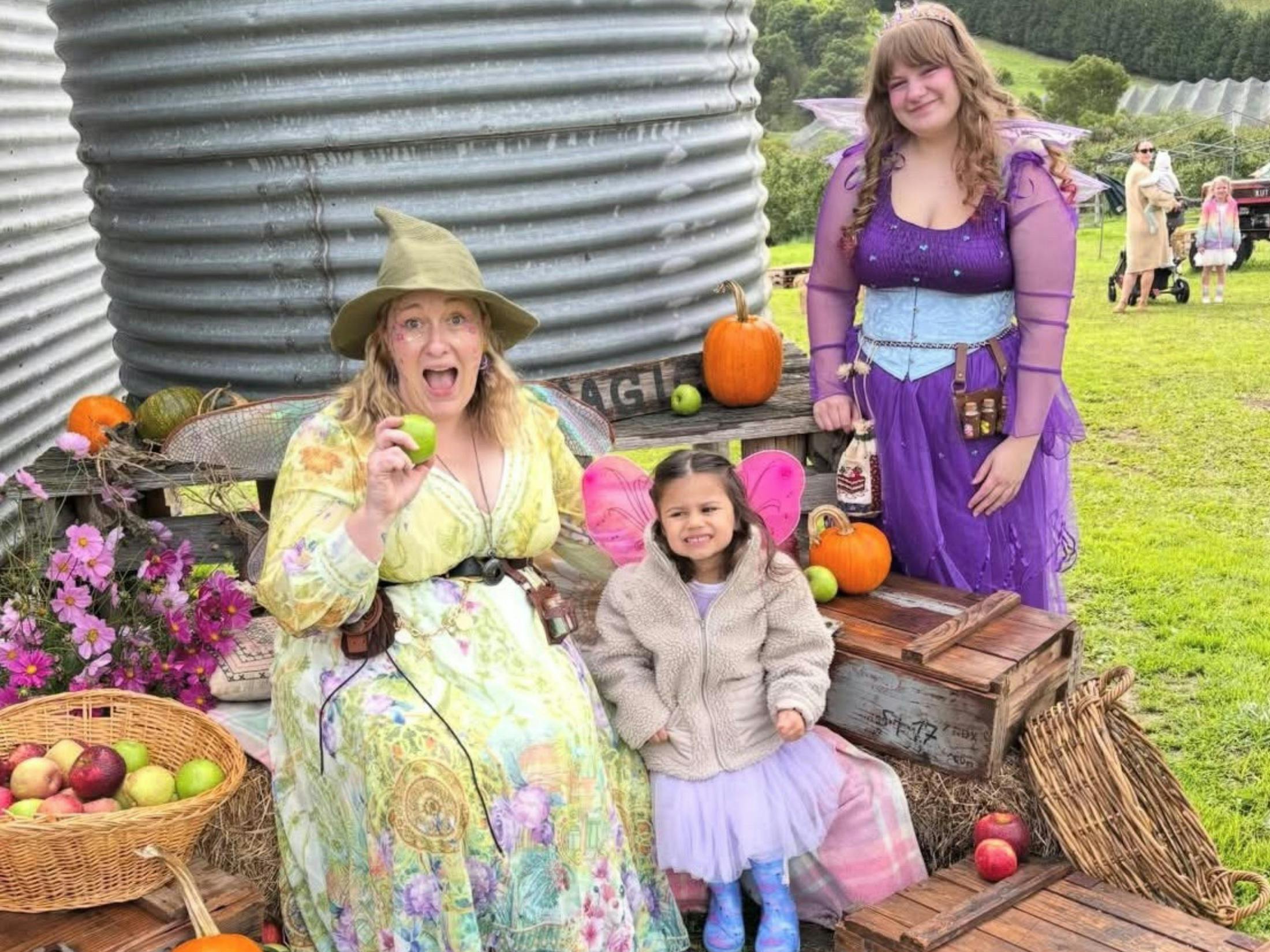 A photo of a girl posing with two fairies surrounded by apples and pumpkins