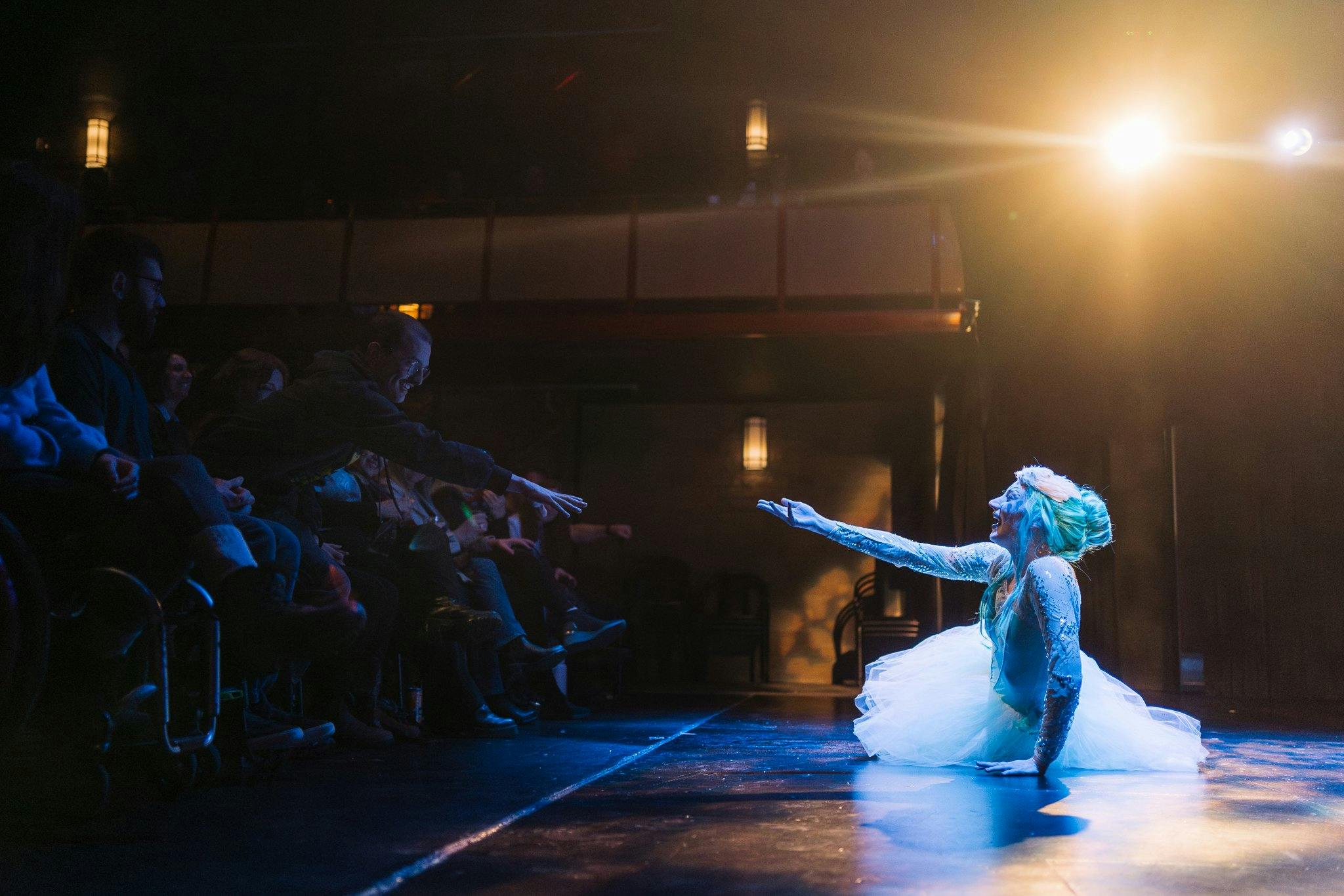 A performer wearing grey skin makeup and a tutu laying on the stage with an arm in the air