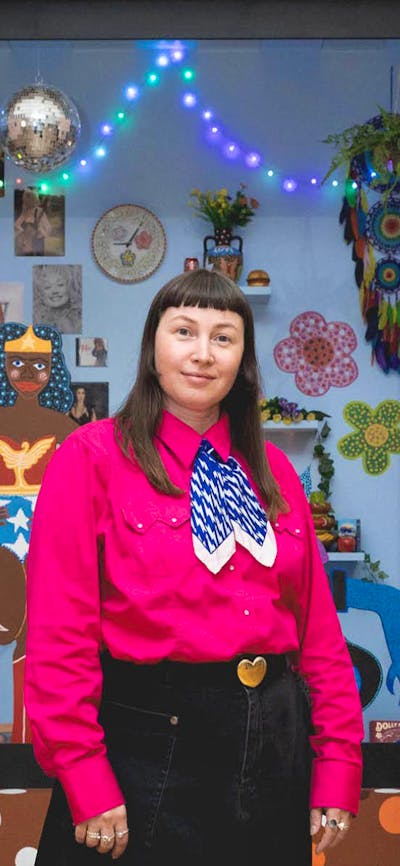 A person wearing a pink shirt stands in front of an 3D artwork featuring a disco ball and figures.