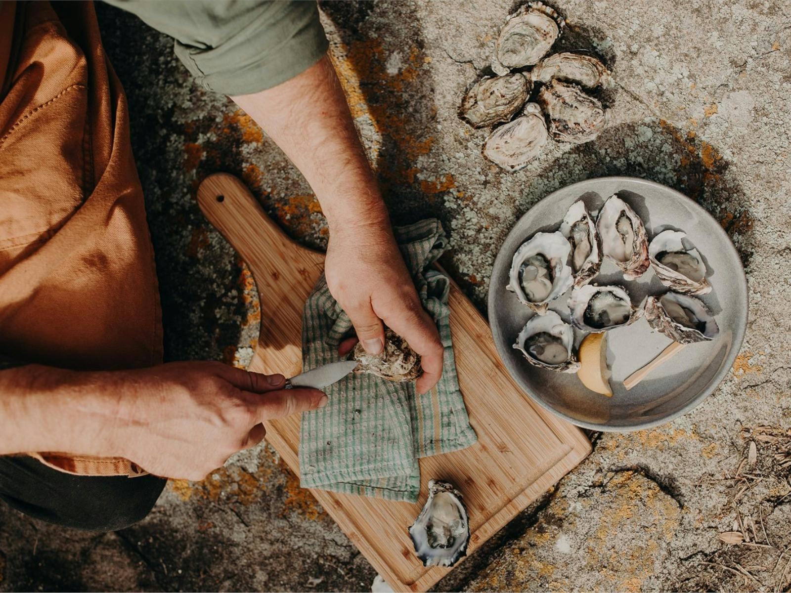 Hands shucking oyster with knife on chopping board. Bowl of fresh oysters and lemon next to board.
