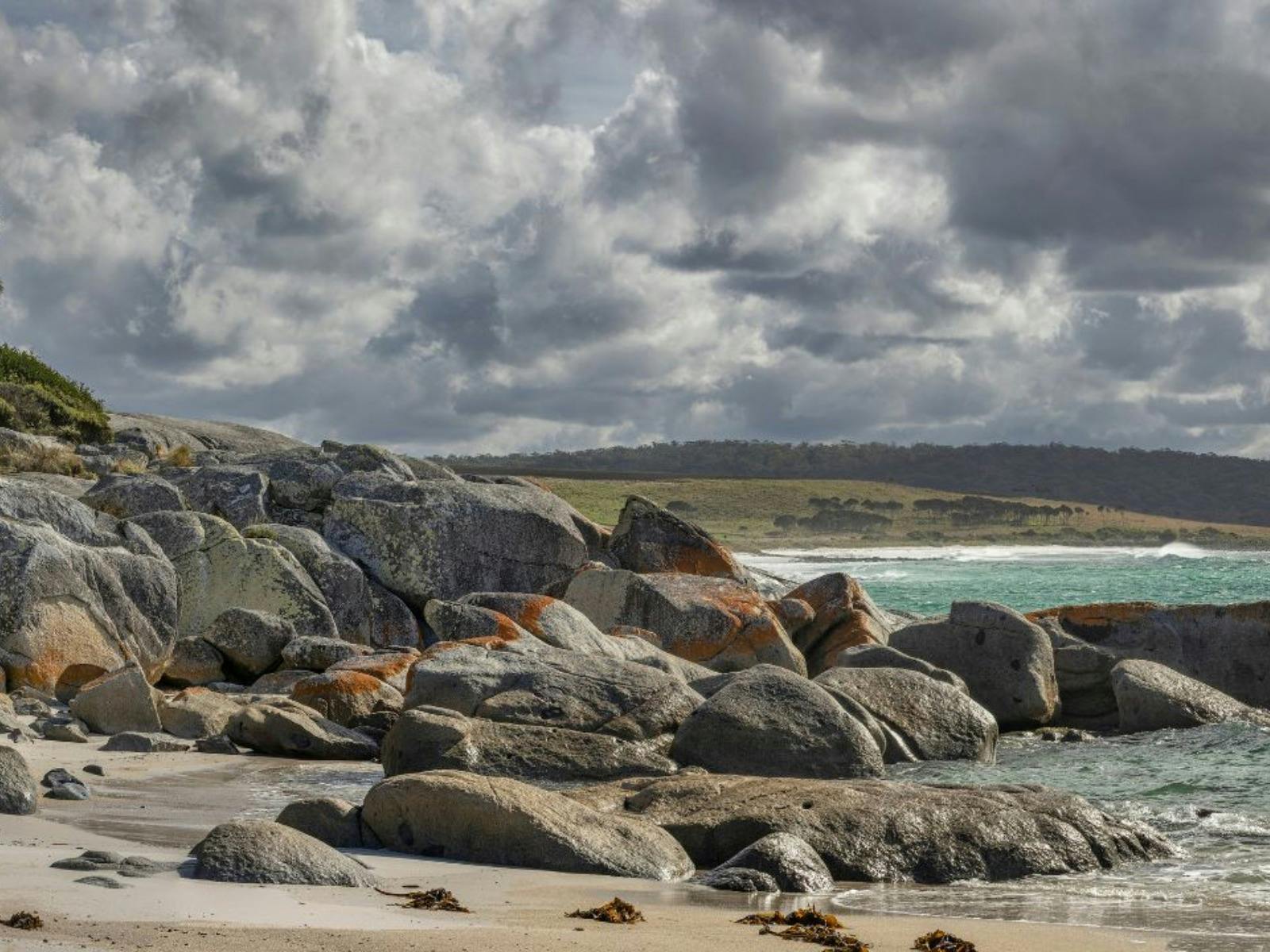 Beach with large grey and orange-stained granite boulders under a cloudy sky.