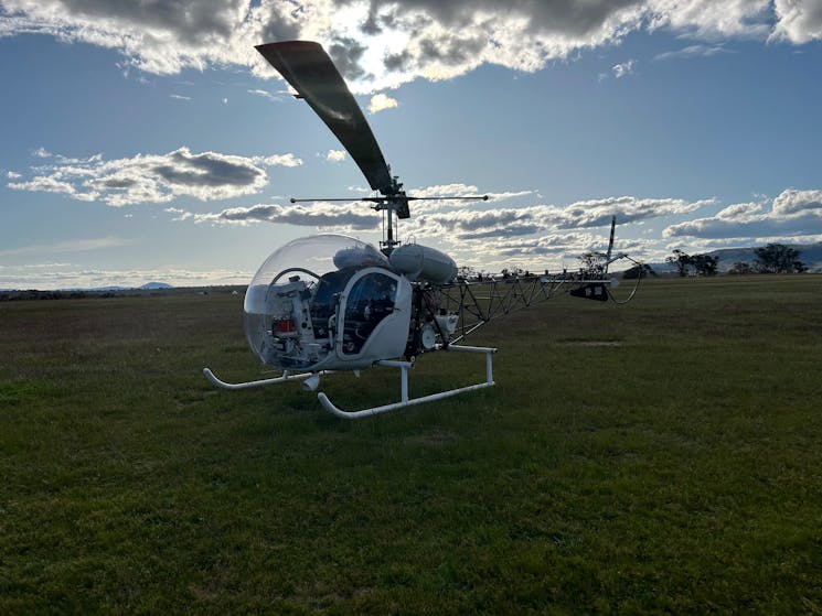 A photo of a Bell 47 helicopter landed in a field, backed by a slightly cloudy blue sky.