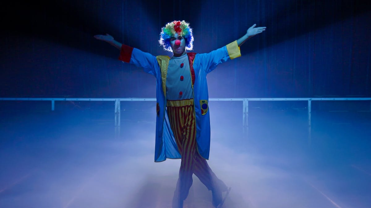 1 clown standing in the middle of the ice skating rink under a spotlight with his arms open wide