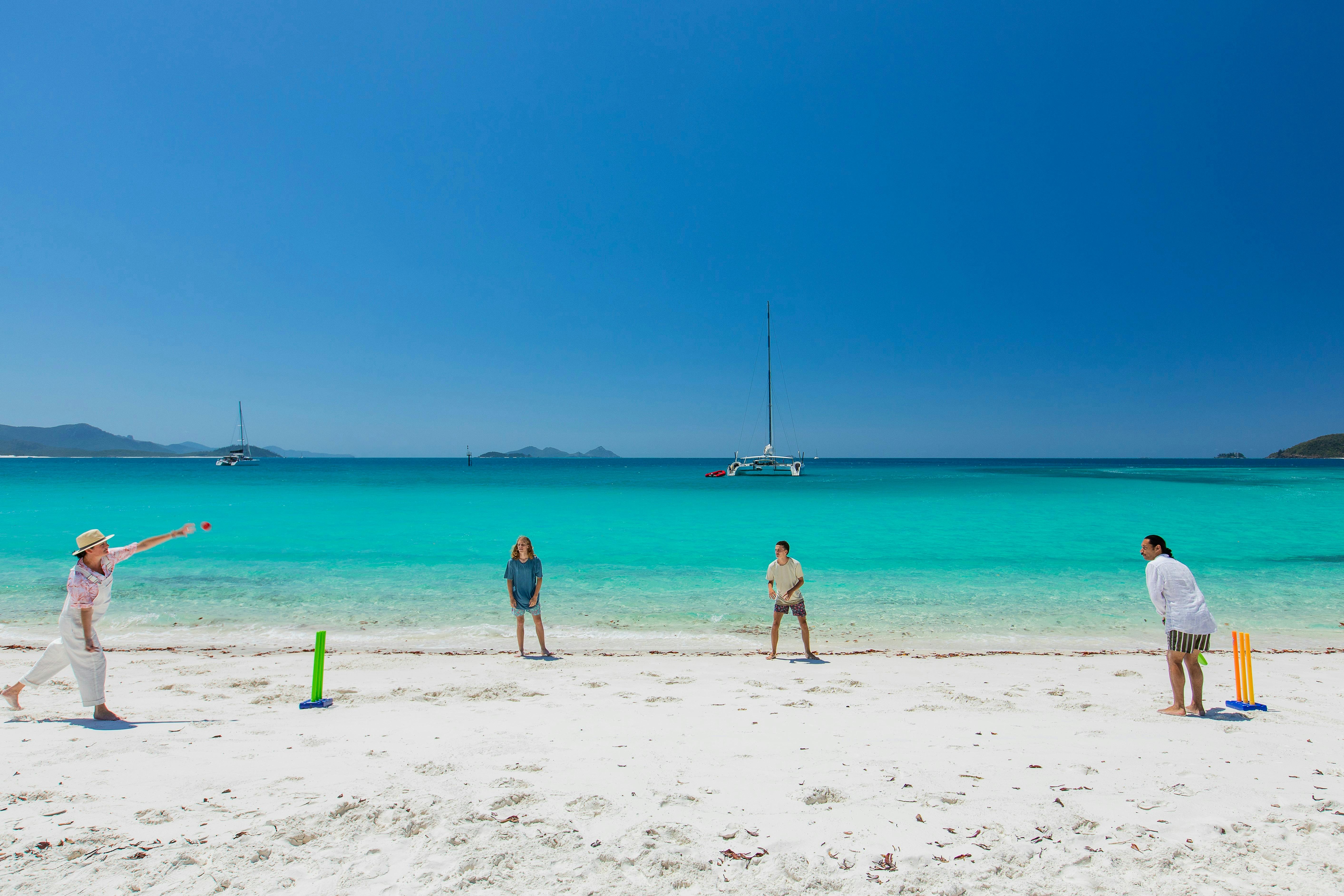 People playing cricket on the white sandy beach with crystal clear ocean and boats in the background