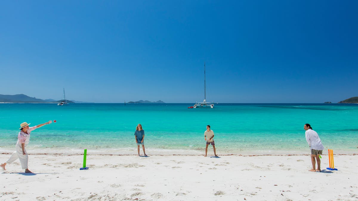 People playing cricket on the white sandy beach with crystal clear ocean and boats in the background