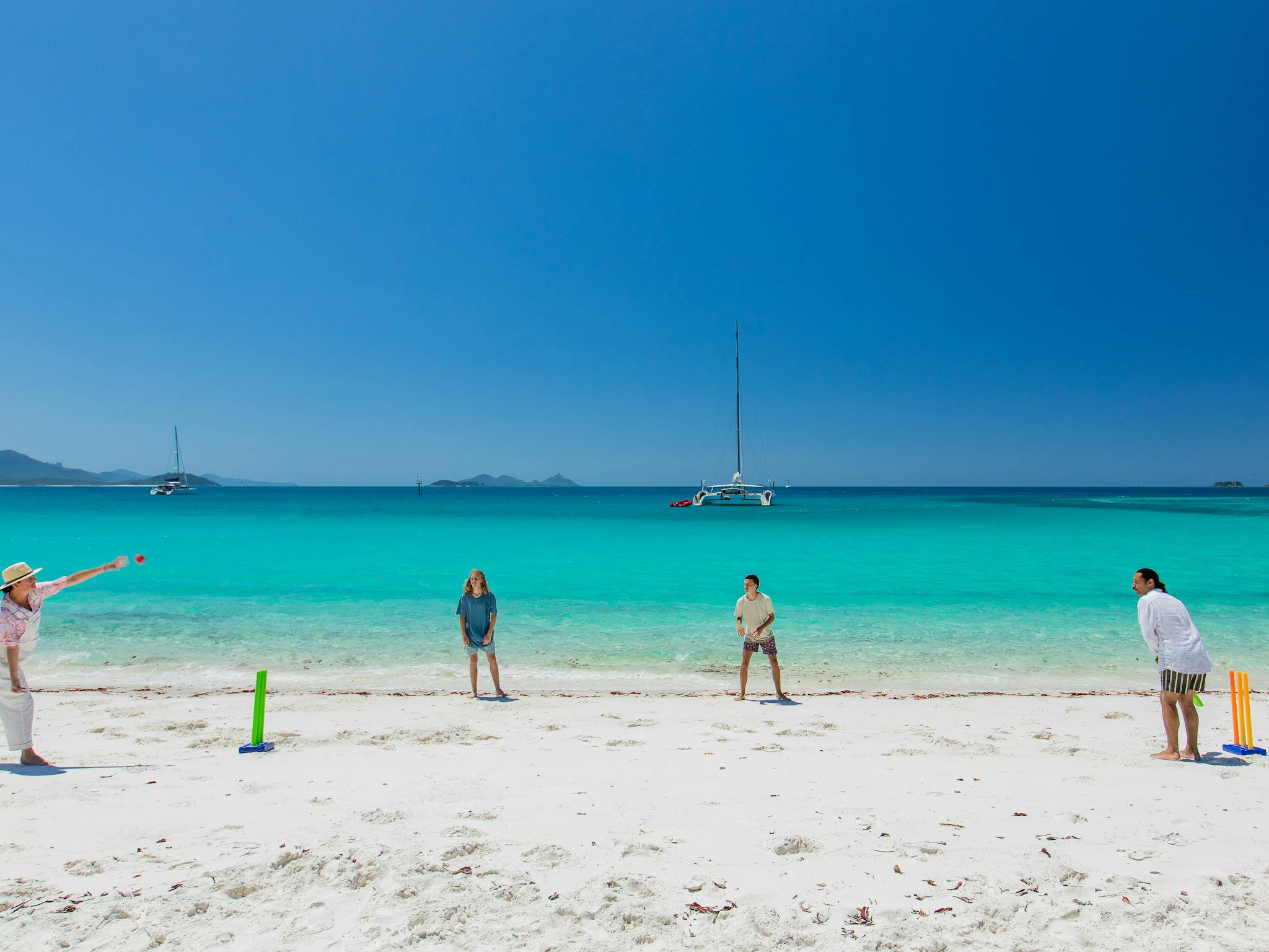 People playing cricket on the white sandy beach with crystal clear ocean and boats in the background