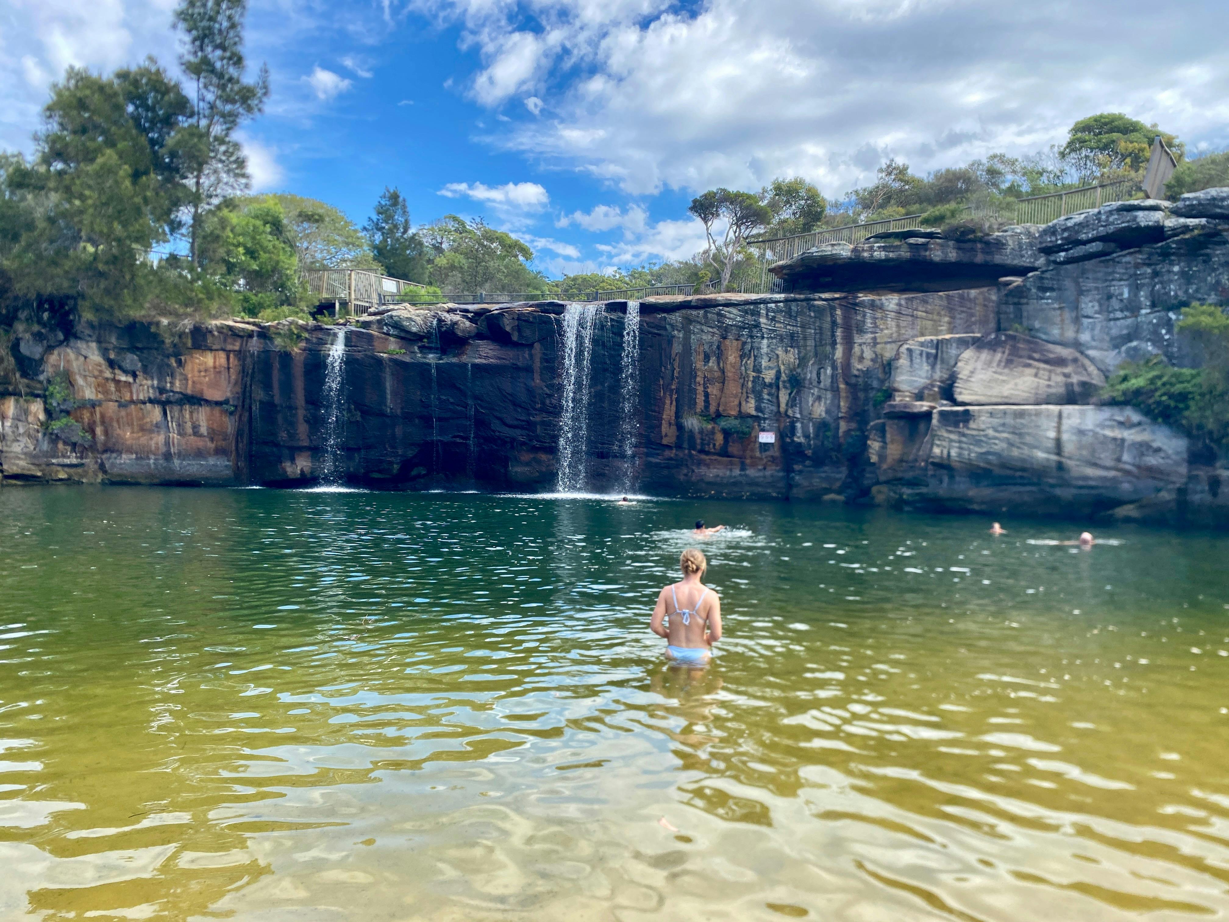 Person swimming in a clear rock pool beneath a waterfall and sandstone cliffs