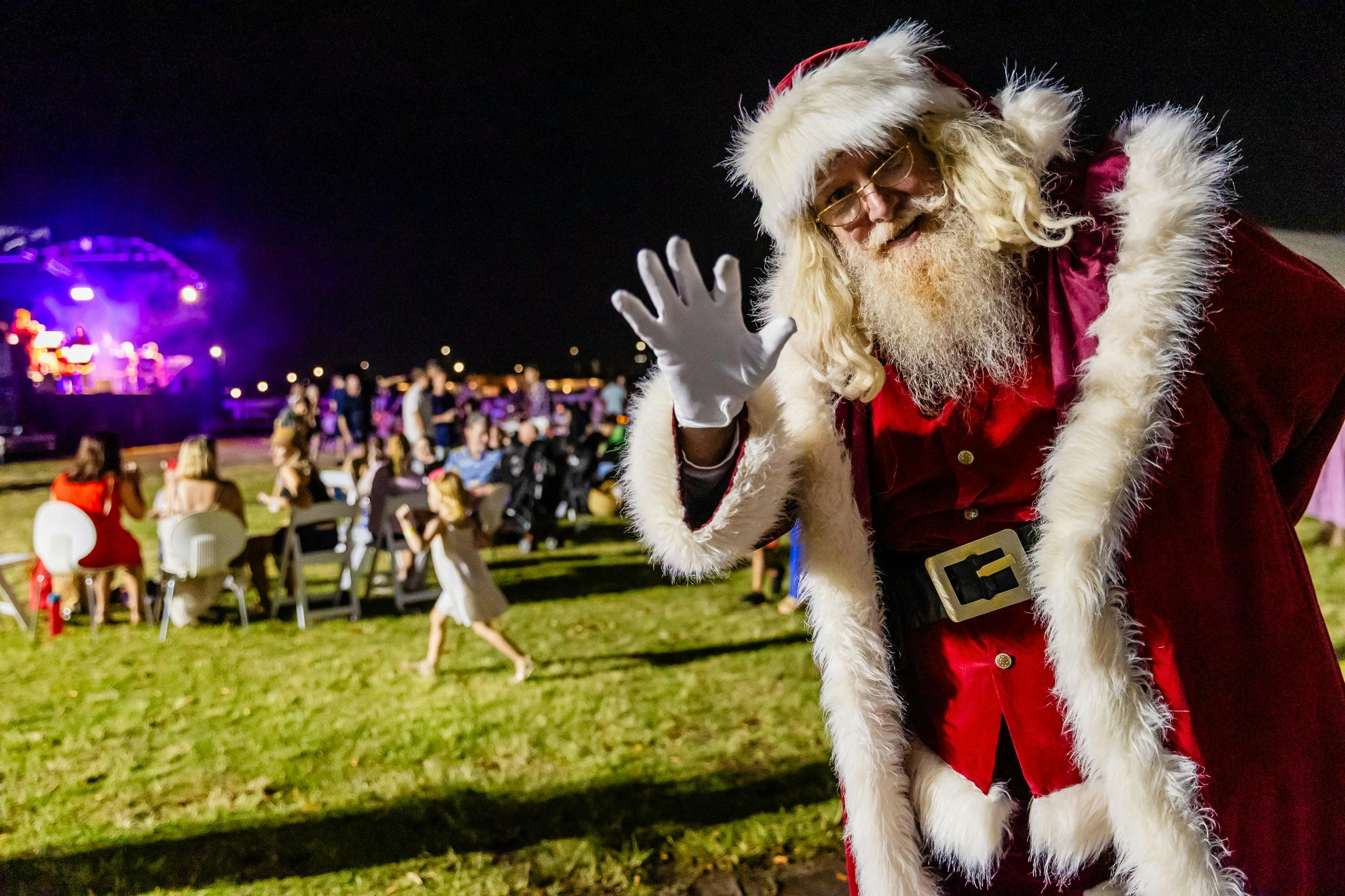 Santa waves to the camera, Christmas carols event takes place behind him