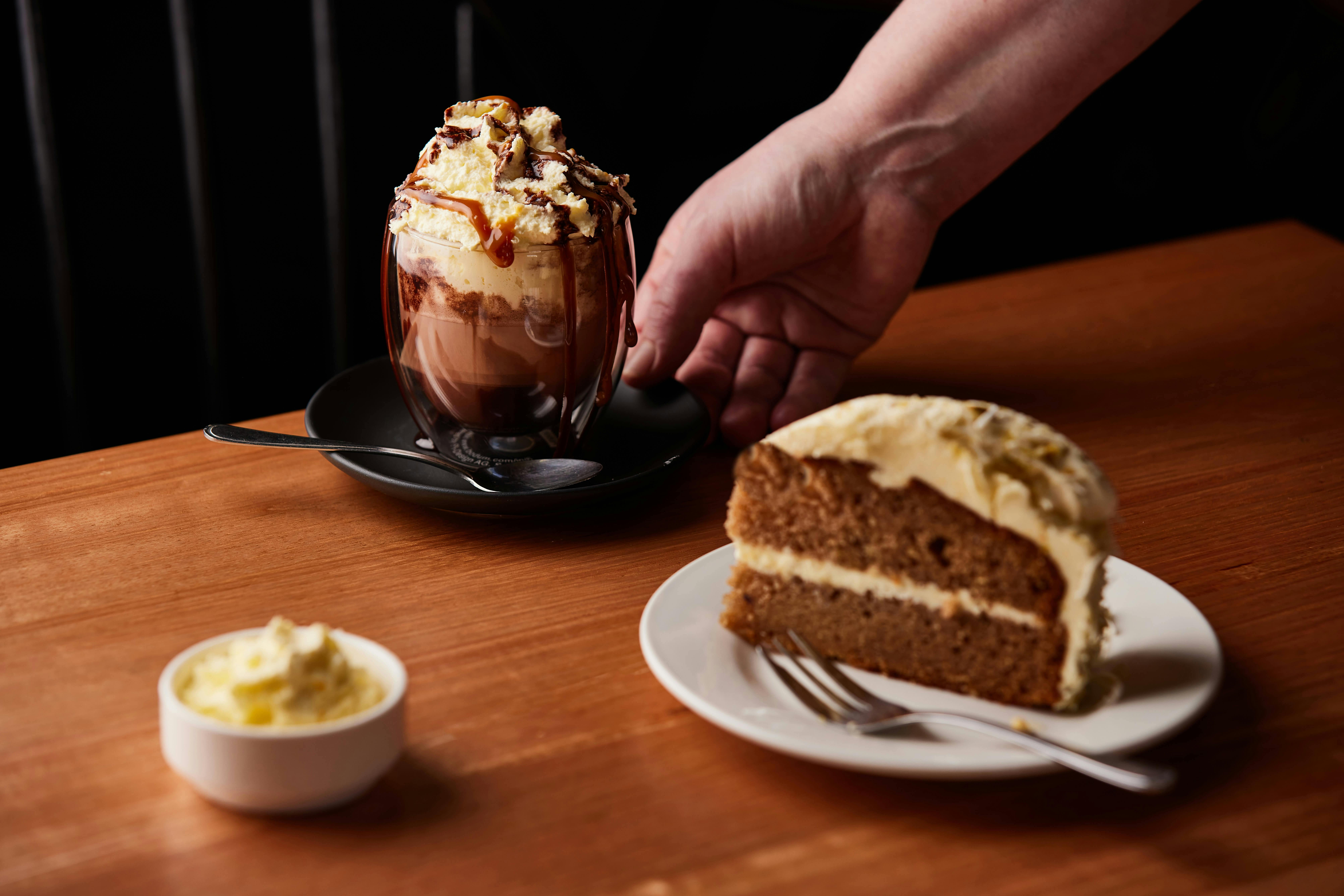 A carrot cakes topped with nuts and cream cheese sits in the foreground with a vienna coffee