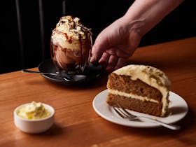 A carrot cakes topped with nuts and cream cheese sits in the foreground with a vienna coffee