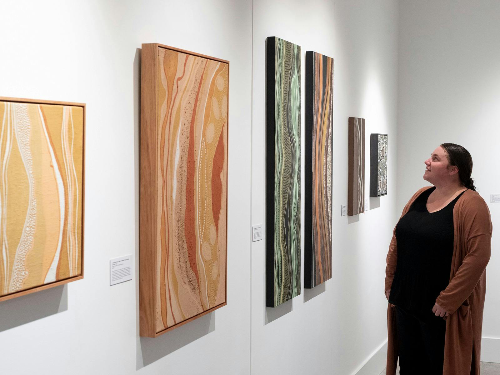 A female Indigenous artist is standing looking at her paintings in the gallery