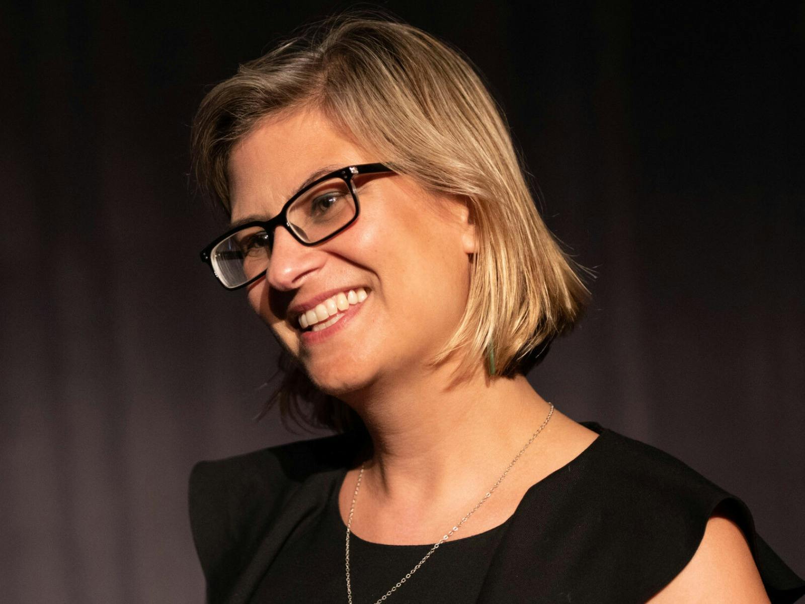 A photograph of Dr Maia Nuku smiling, wearing black-framed glasses and a black top.