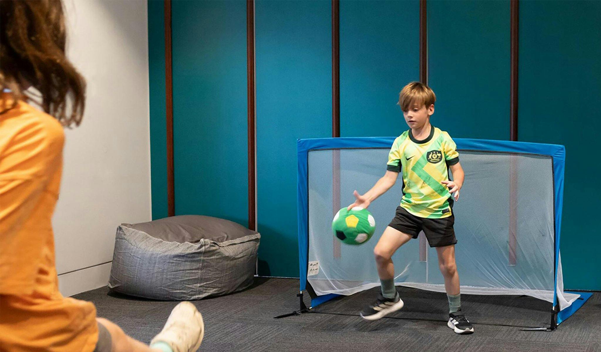 A child kicks a soccer ball towards a goal, where a child in a Matildas Jersey defends the ball.