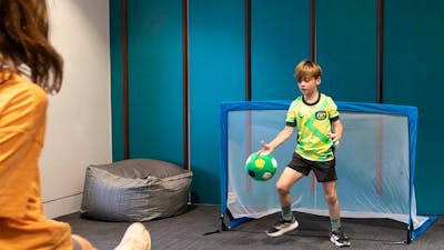 A child kicks a soccer ball towards a goal, where a child in a Matildas Jersey defends the ball.