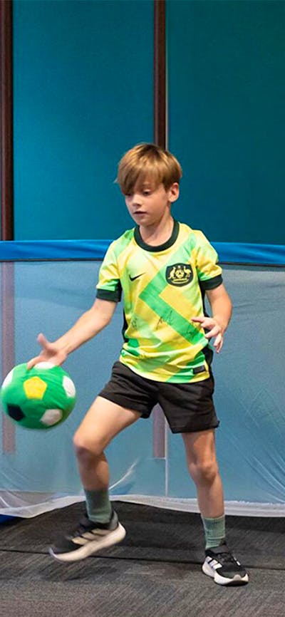 A child kicks a soccer ball towards a goal, where a child in a Matildas Jersey defends the ball.
