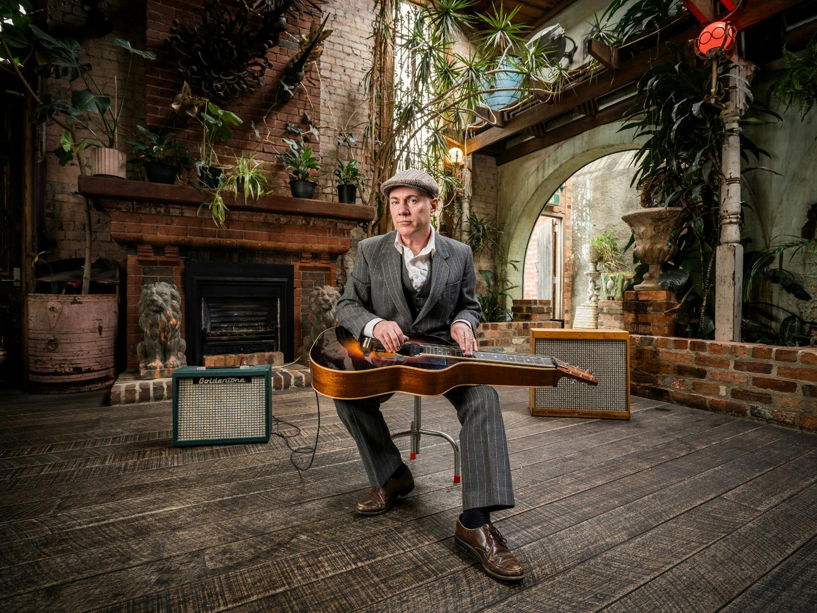 A person in a gray suit and flat cap sits on a stool in front of a fireplace, with a vintage guitar