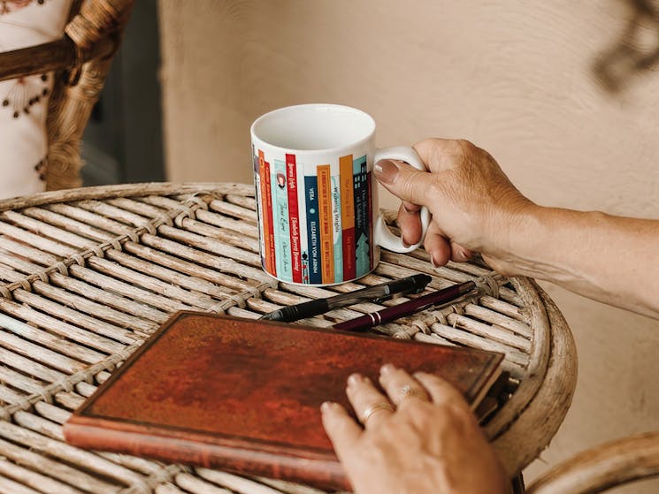 A small round table with a writing journal and pens a hand is resting on a coffee cup