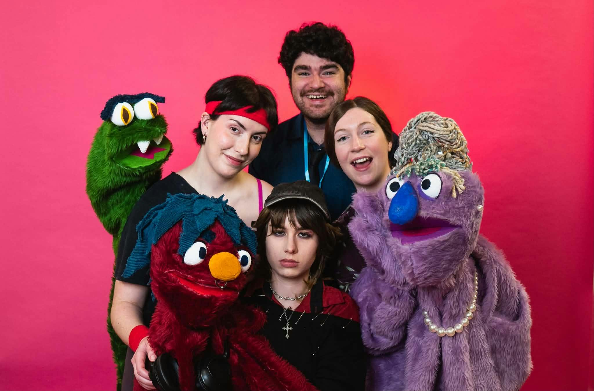 4 humans holding 3 puppets near them. They are red, Green and Purple sweet monster looking puppets