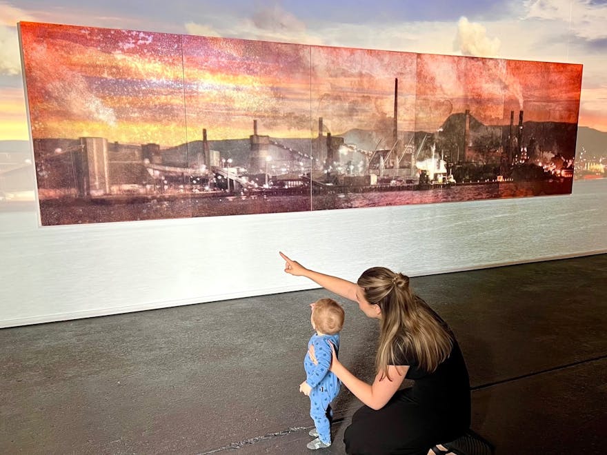 a woman and toddler looking at a Painting of Port Kembla Steelworks with moving image overlay
