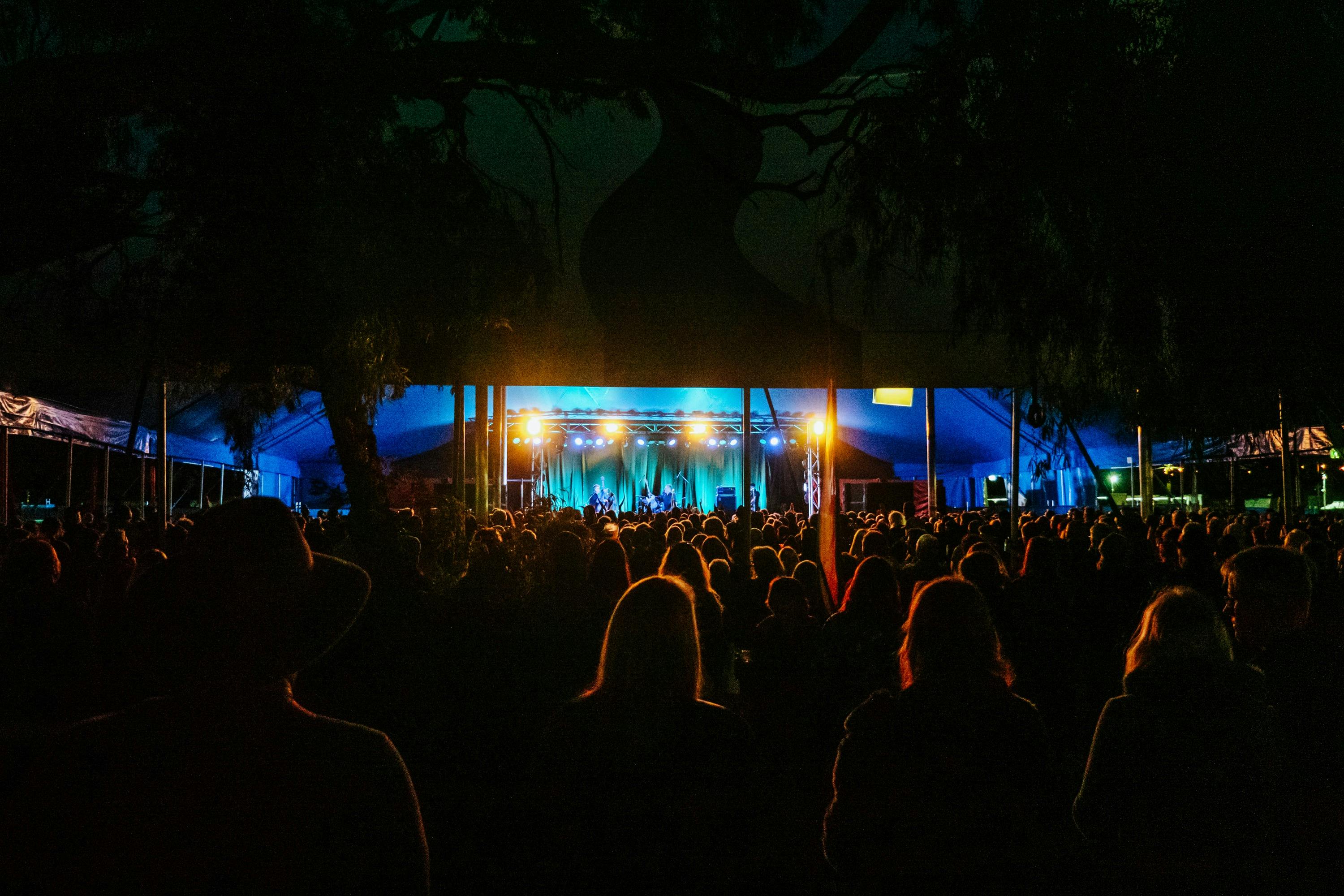 Night time crowd watching band on stage under lights, inside a circus tent.
