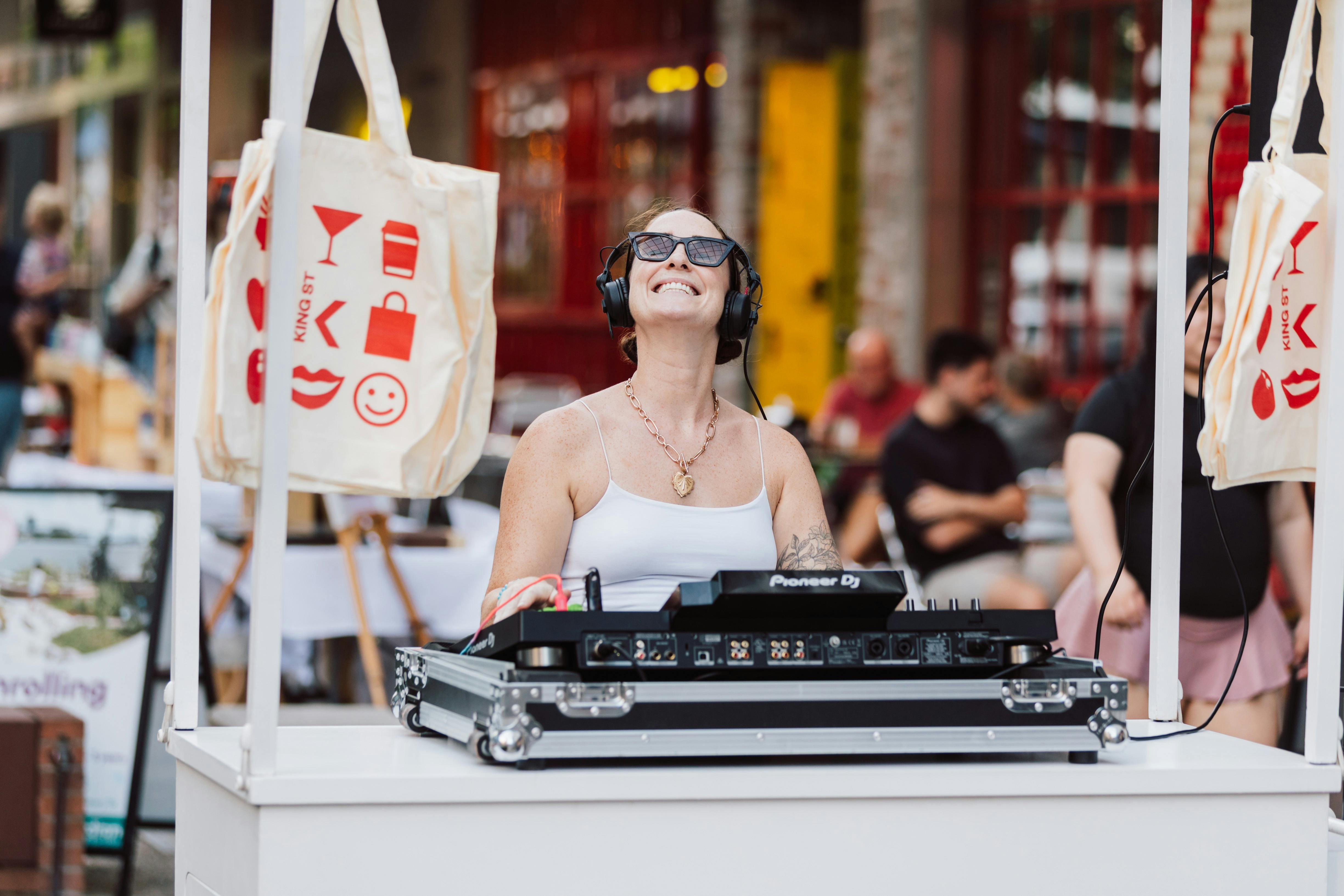 Smiling DJ wearing headphones and sunglasses performs at an outdoor market stall.