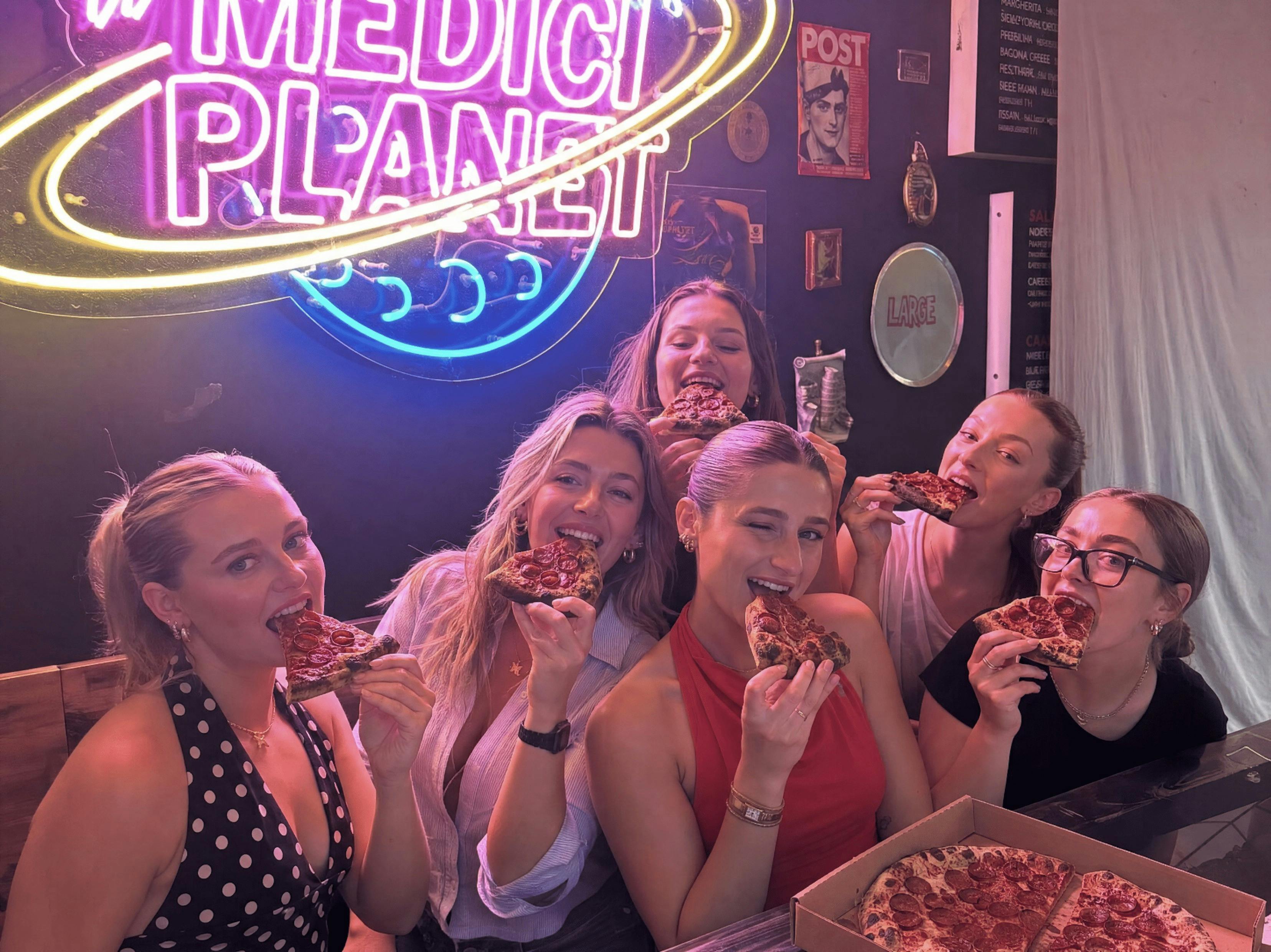 Six medici staff eating pizza slices under a neon sign      inside Medici Pizzeria