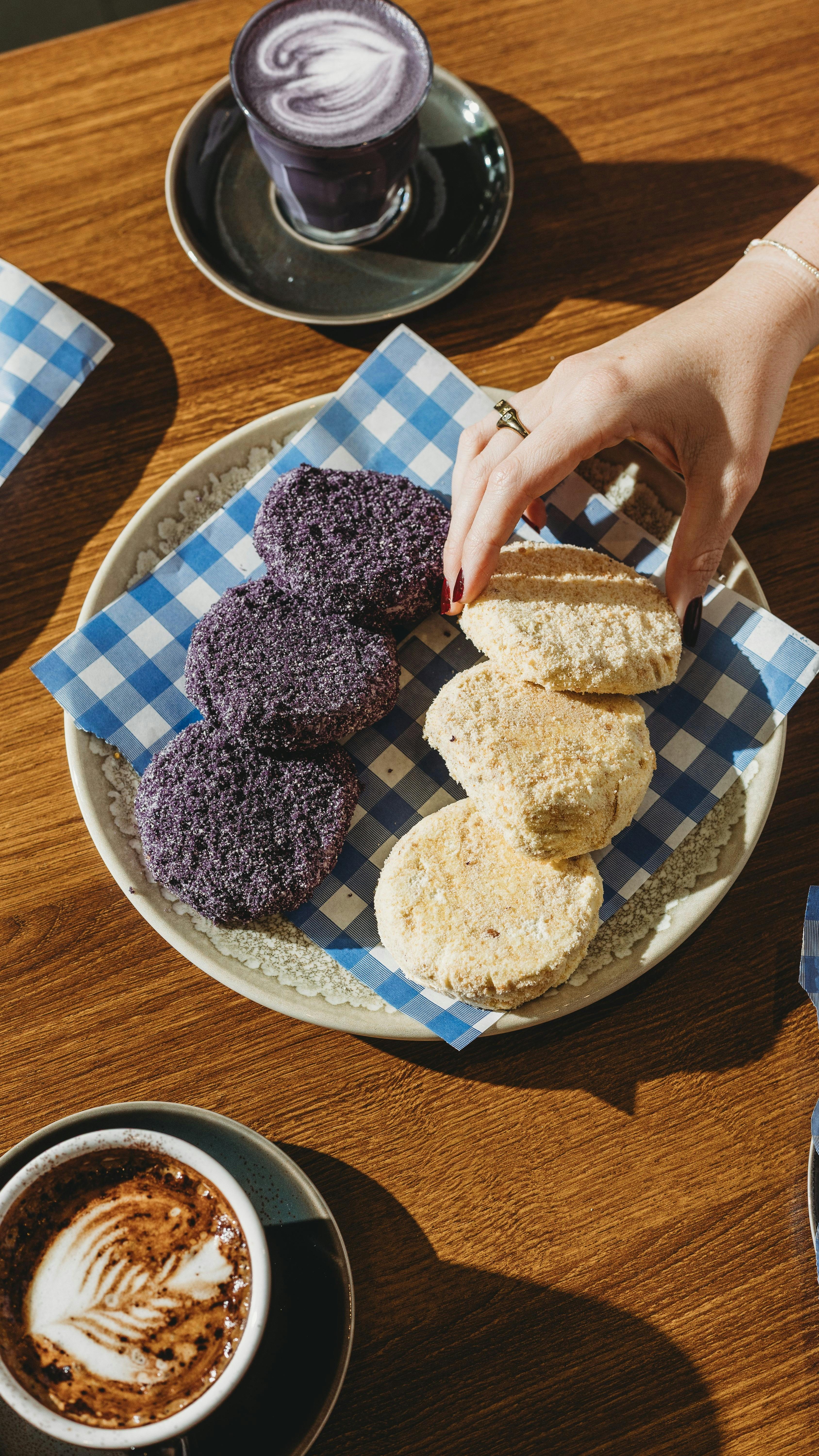 A plate of purple and beige pastries on checkered paper, placed on a wooden table with two cups of c