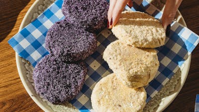 A plate of purple and beige pastries on checkered paper, placed on a wooden table with two cups of c
