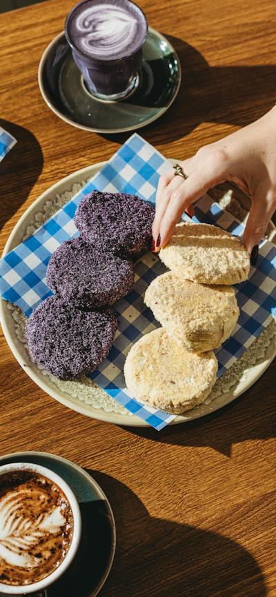 A plate of purple and beige pastries on checkered paper, placed on a wooden table with two cups of c