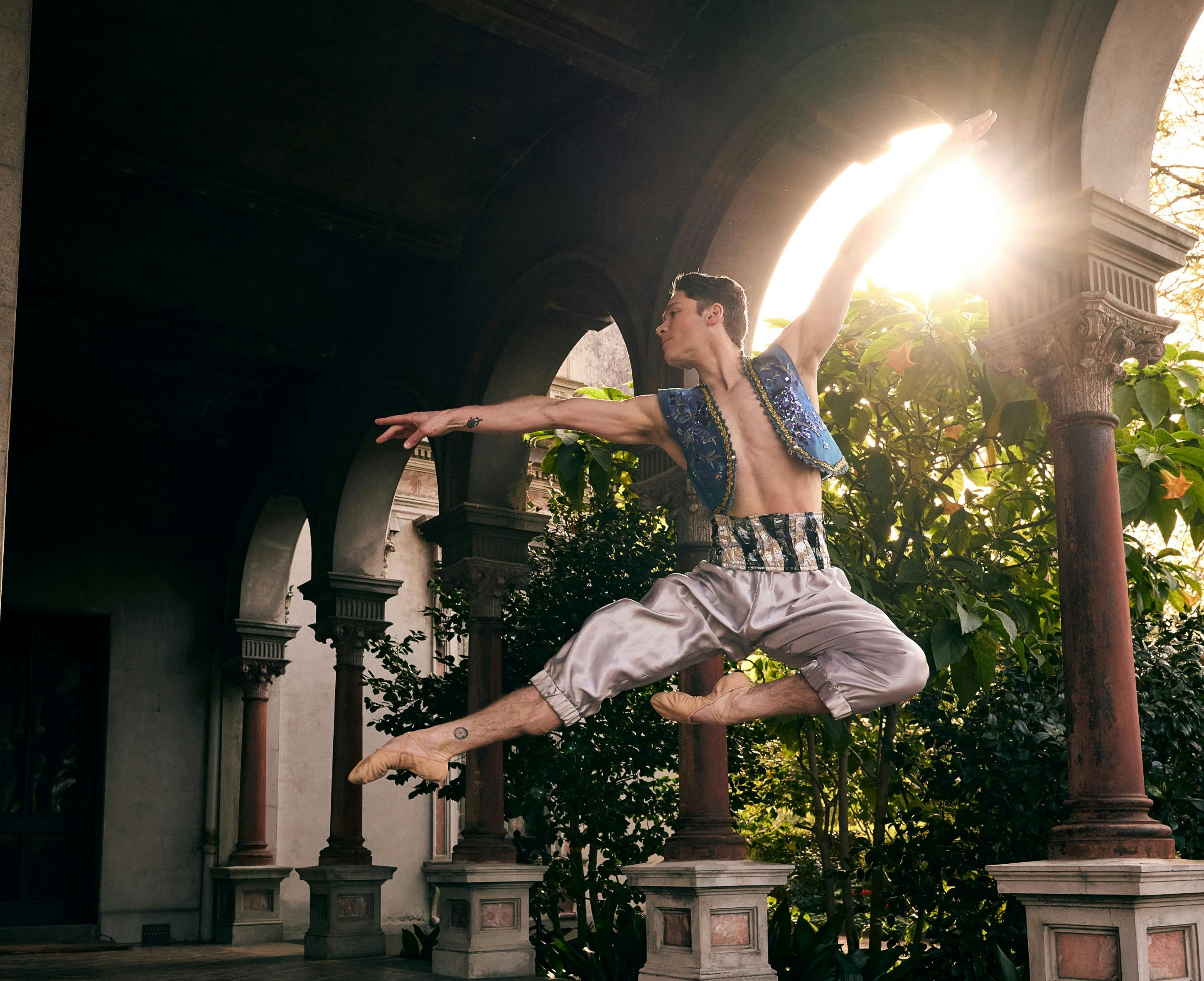 Ballet dancer leaps in ornate costume beneath stone arches, sunlight behind.