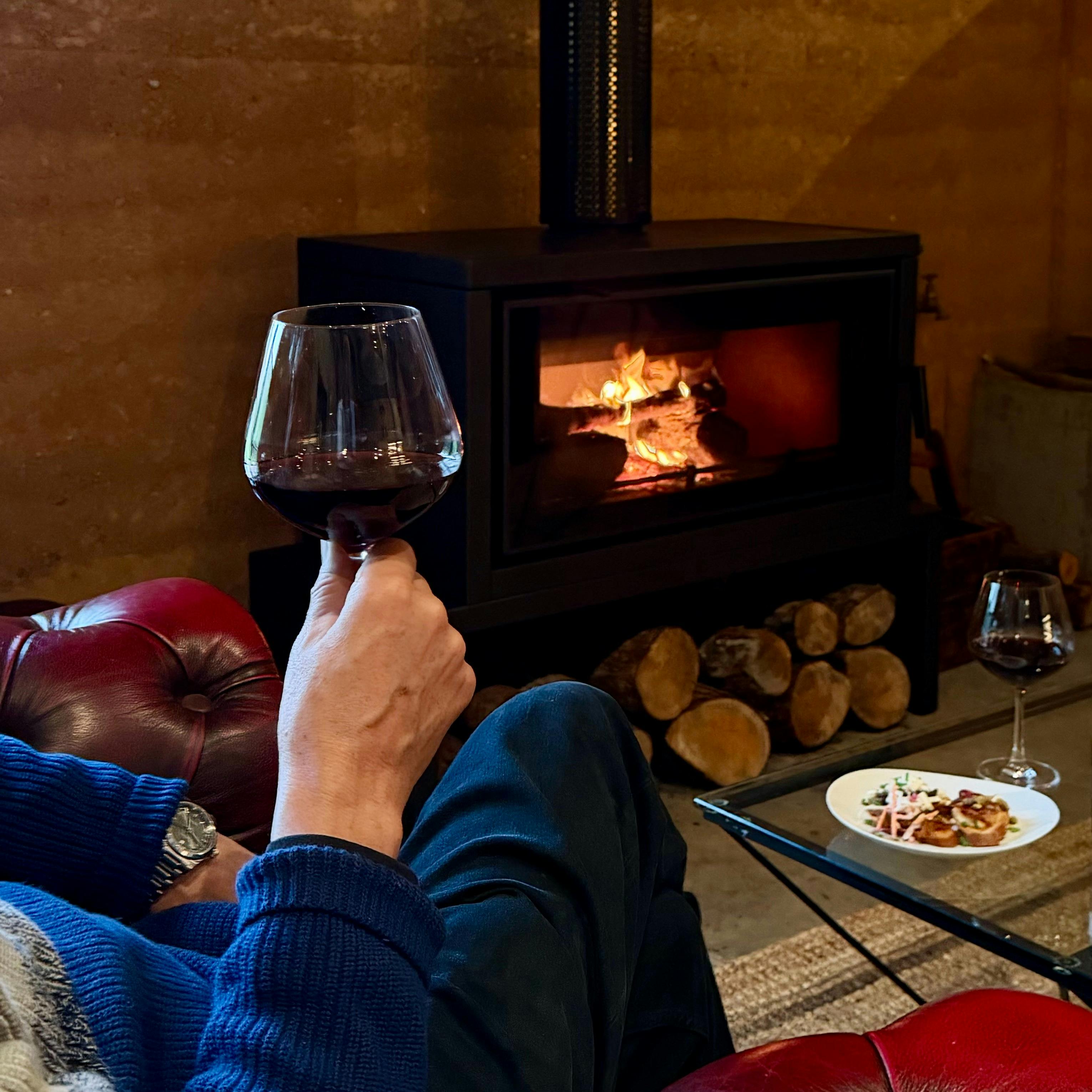 Somebody sitting in an armchair by a woodburner drinking a glass of red wine