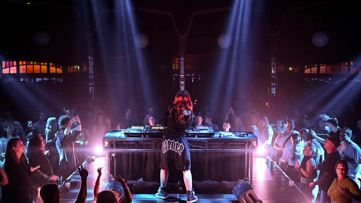 A woman playing electronic DJ equipment on a stage with a crowd dancing around her.