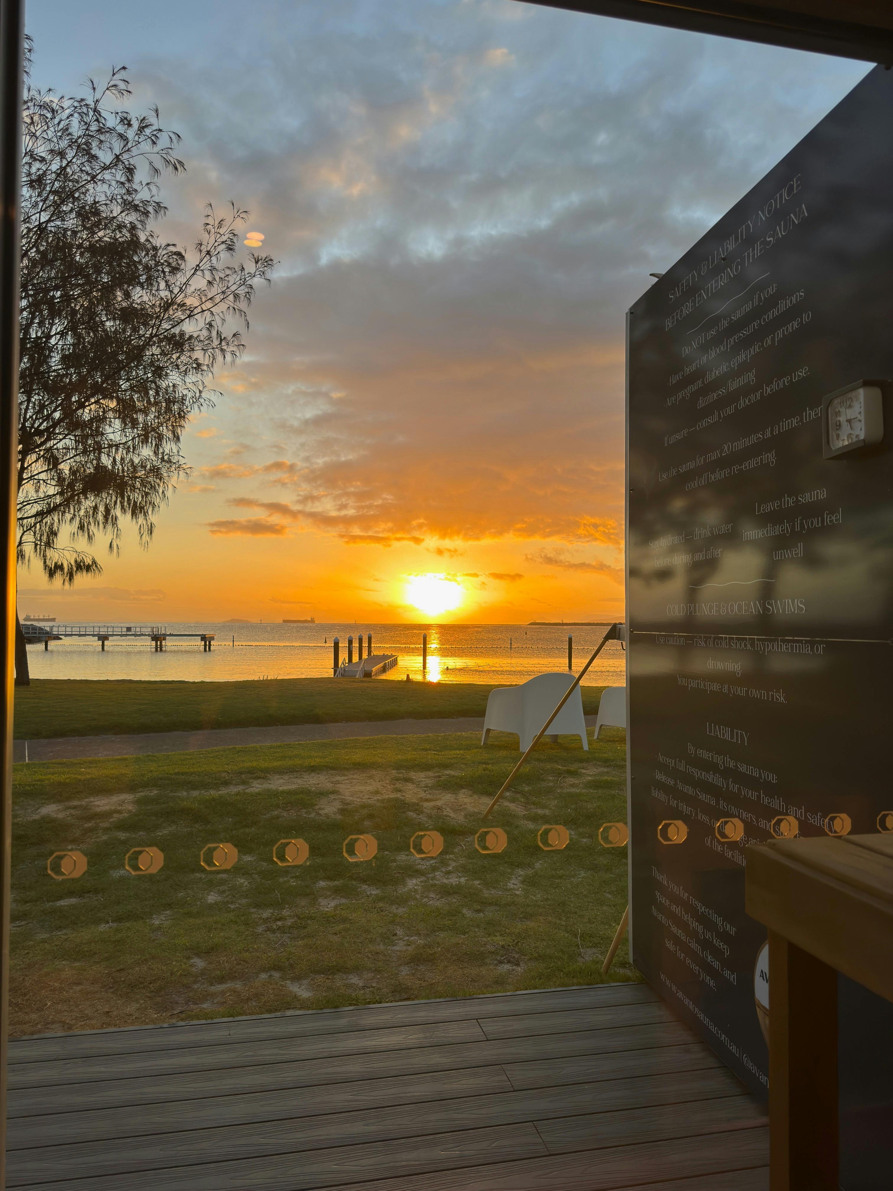 Golden sunrise over still ocean viewed from inside sauna, jetty and chairs silhouetted.